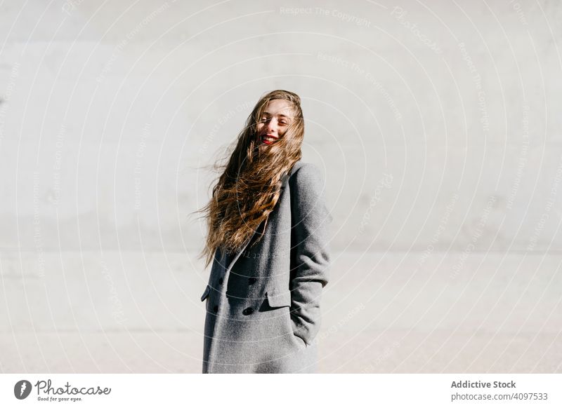 Young woman in coat on windy day stylish street closed eyes wall city building female urban fashion cool young model outfit warm weather exterior lady long hair