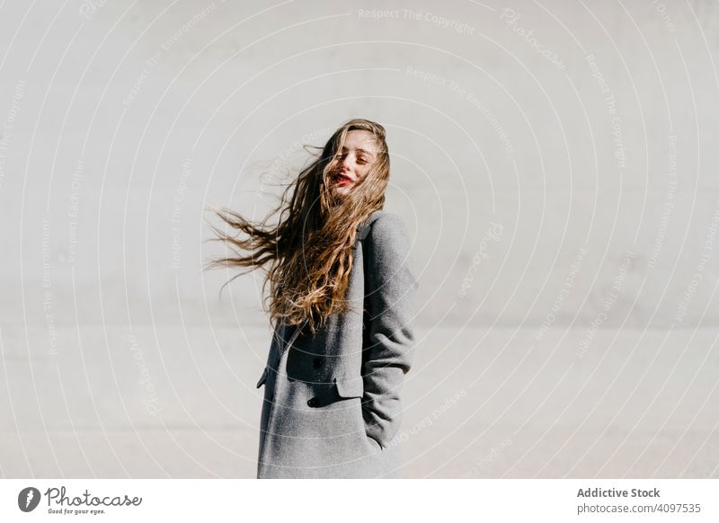 Young woman in coat on windy day stylish street closed eyes wall city building female urban fashion cool young model outfit warm weather exterior lady long hair