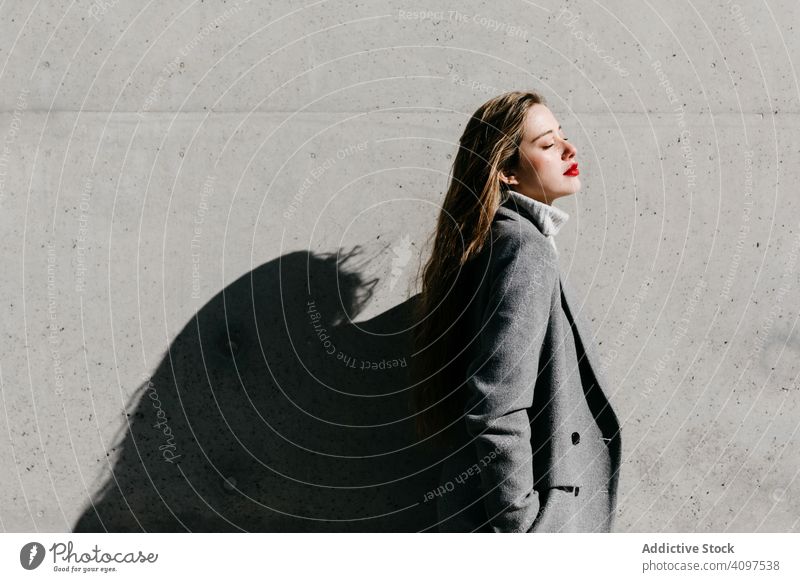 Young woman in coat on windy day stylish street closed eyes wall city building female urban fashion cool young model outfit warm weather exterior lady long hair