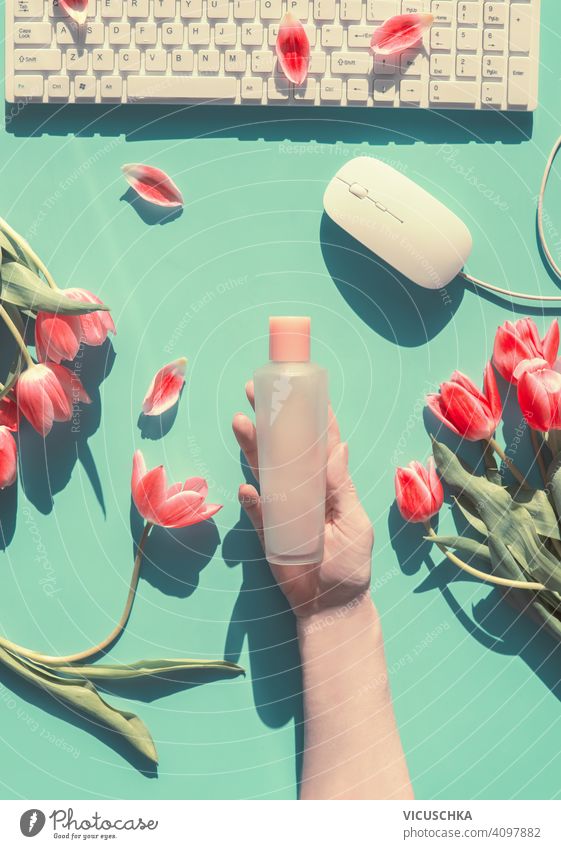 Woman hand holding cosmetic bottle on green desk background with pink tulips, white computer keyboard and mouse. Springtime home office. Natural cosmetic woman