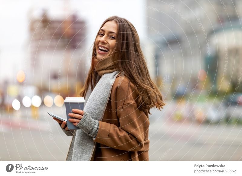 Smiling young woman with smart phone and coffee cup outdoors at urban setting autumn fall girl female beautiful weather coat fashion pretty attractive adult