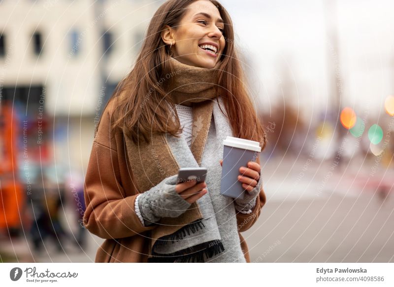 Smiling young woman with smart phone and coffee cup outdoors at urban setting autumn fall girl female beautiful weather coat fashion pretty attractive adult