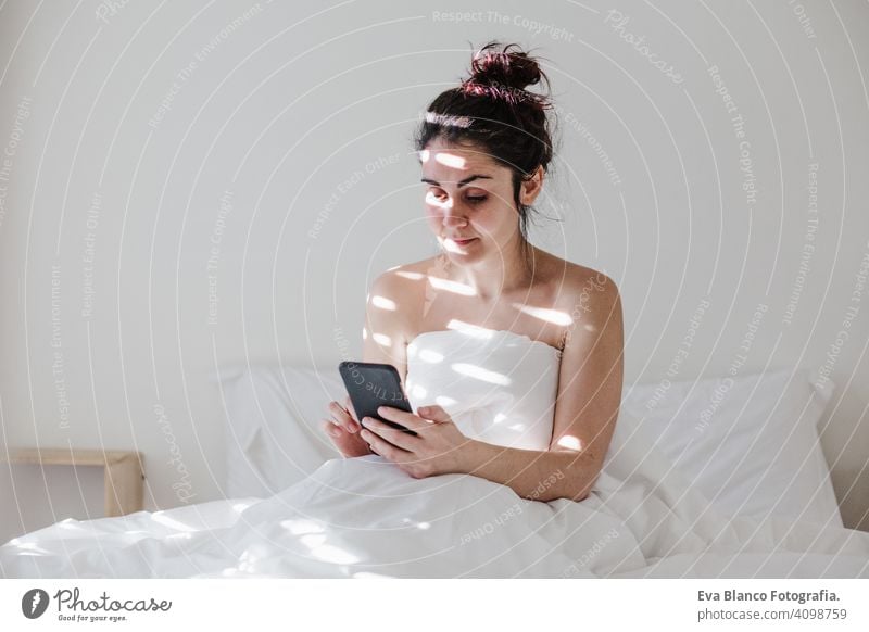 attractive young caucasian woman relaxing well in bed during morning time. Lady using mobile phone in bedroom technology white sheets arousing call talking