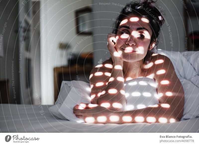 portrait of attractive young caucasian woman relaxing in bed during morning time. Lady enjoys fresh soft bedding linen and mattress in bedroom. Blind shadows on face