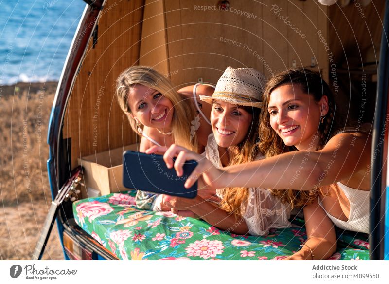 Relaxed girl friends taking selfie comfortable placing on trunk of blue car women smartphone hugging using travel minivan having fun gadget device ocean hippie