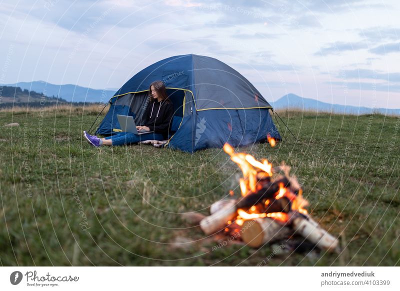 Girl with laptop in the mountains. Tourist woman uses a laptop in nature, freelancer typing on a computer near a tent in a journey against the backdrop of a mountain landscape.