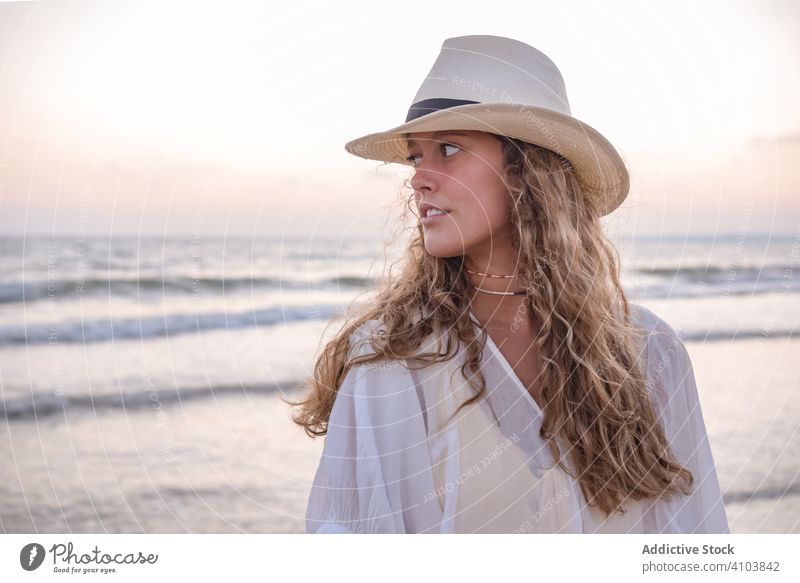 Charming woman in light white dress on wavy beach sea traveling seaside tourism charming vacation hat curly holiday summer freedom journey young relaxing