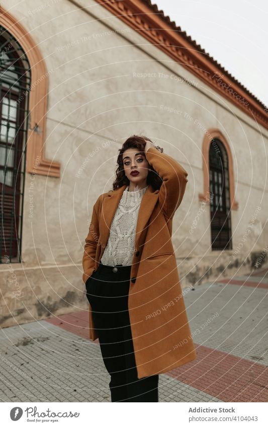 Young woman in overcoat with arms raised looking at camera with provocative eyes young standing curly haired challenge jumper knitted building warm female