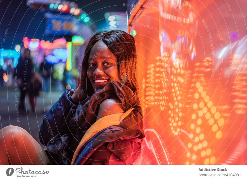 Cheerful black woman on funfair smile night arcade illumination sit rest weekend ethnic female amusement attraction colorful joy happy cheerful bright lady