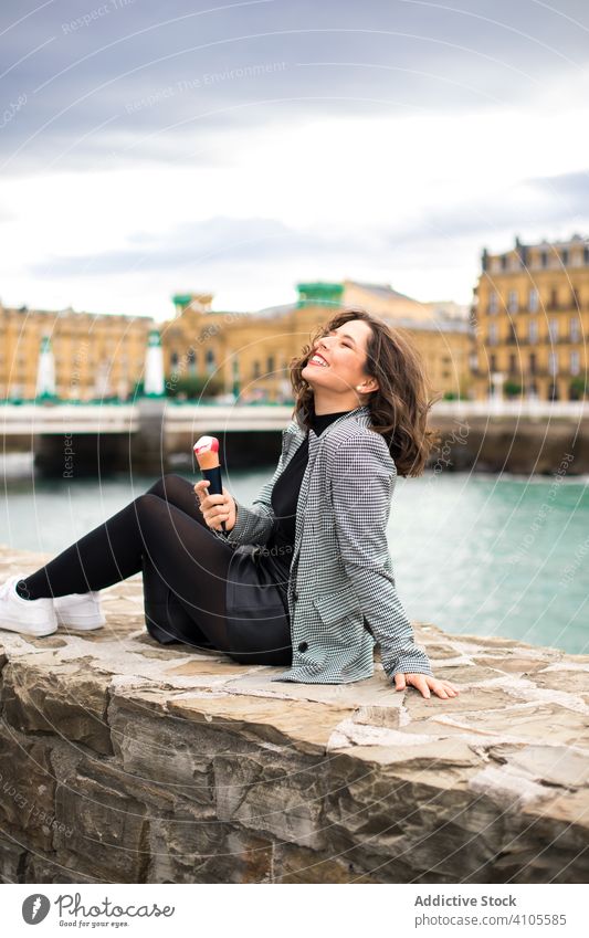 Satisfied woman enjoying tasty ice cream cone at quay smile laugh sitting fence waterfront female delicious overcast city street casual positive relax rest