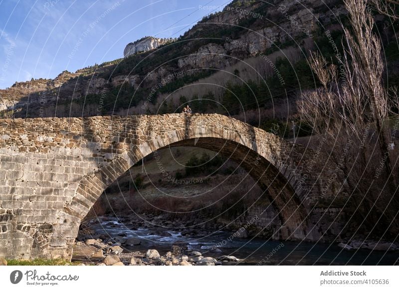 People standing on old bridge above small river in mountain valley forest ancient structure architecture water rocky landscape stone travel nature tourism