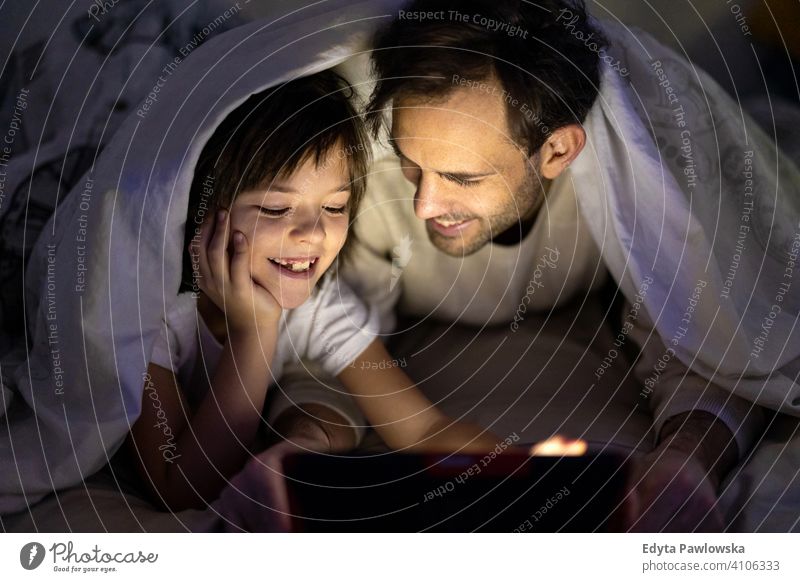 Father and son using digital tablet together at night in bed bedtime lying down reading bedroom under blanket storytelling internet online cute evening sleeping