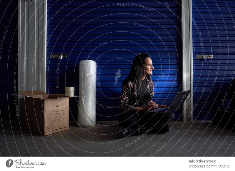 Beautiful brunette woman with plaid shirt and black pants sitting in front of the door of a storage room. He has a portal computer with which he works and organizes the moving boxes