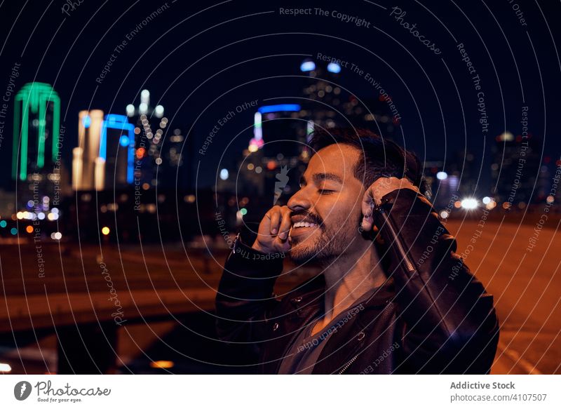 Overjoyed Hispanic guy using headphones during evening walk man smile enjoy city street dusk night jacket black casual device gadget ethnic hispanic latino