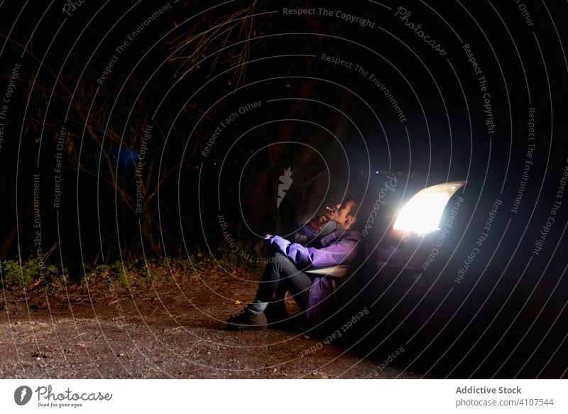 Cool young crouched woman smoking cigarette beside car on road at twilight night cool dark headlight smoke hipster fire thoughtful confident bad habit