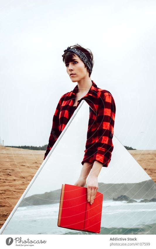 Woman with mirror and book standing on beach woman concept melancholy solitude thoughtful red reflection sea serious pensive casual young female ocean sand