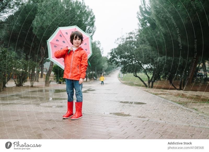 Cute child with colorful umbrella looking at camera in park alley rain season funny water wet dirt childhood mud autumn game active weather rubber boots