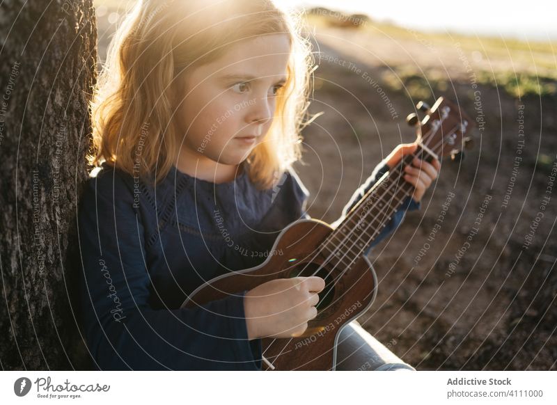 Little girl sitting under tree and playing ukulele kid music guitar nature child summer serious little instrument sound song melody trunk perform practice rest