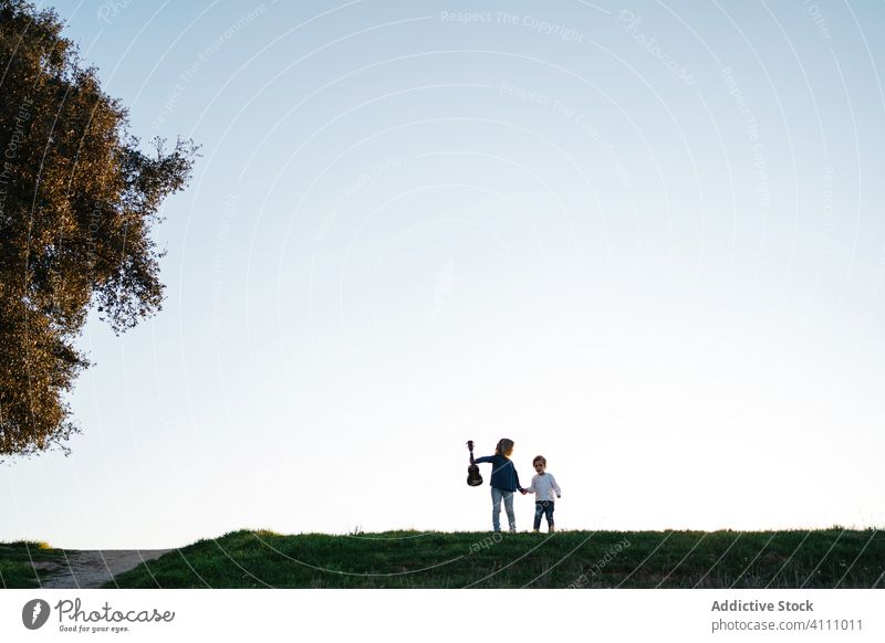 Little sister and brother walking in field children nature summer sunset evening together explore grass ukulele guitar follow sibling friend sky calm tranquil