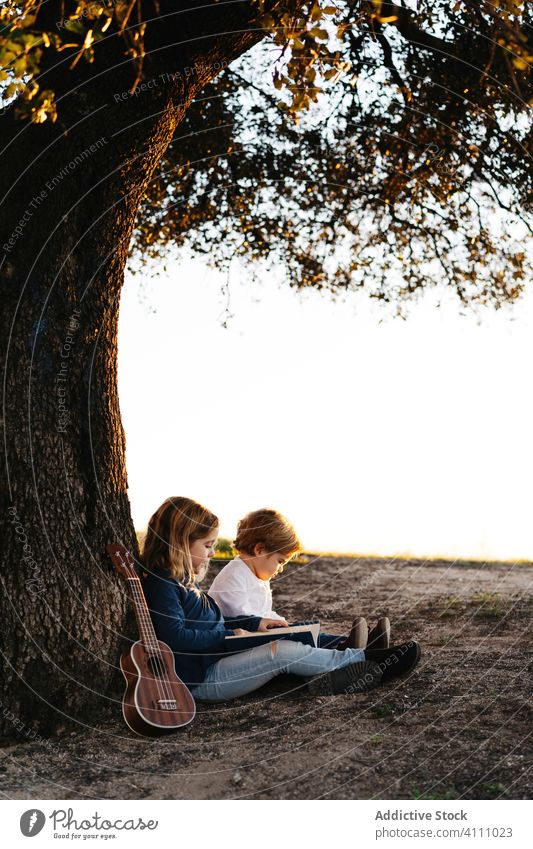 Kids sitting under tree and reading book children nature together summer kid ukulele guitar rest tranquil sister brother sibling friend countryside idyllic