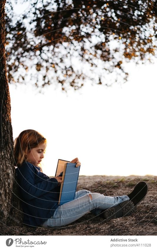 Little girl with book sitting under tree kid read nature field trunk summer casual child calm tranquil serious concentrate female learn meadow lifestyle