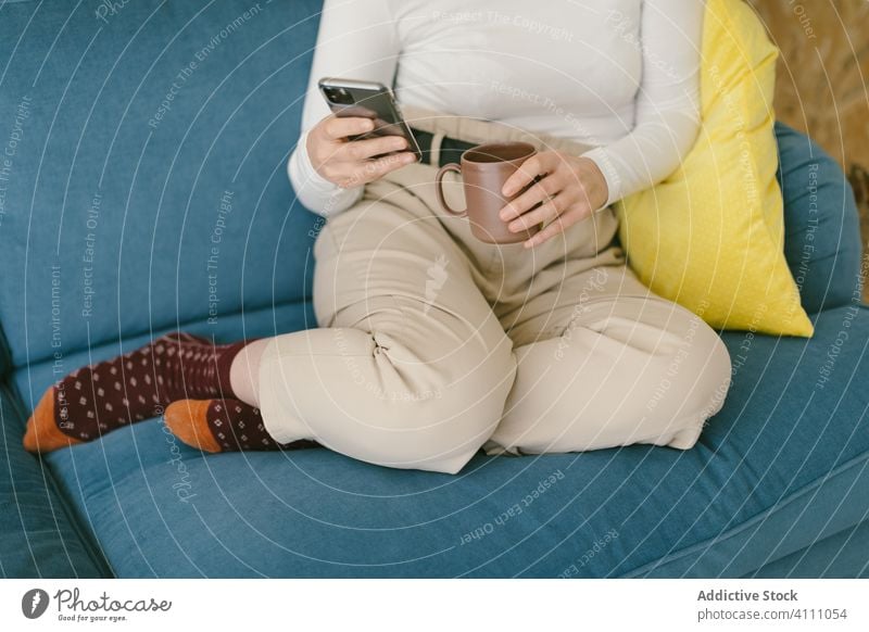 Crop businesswoman resting on couch with coffee and phone break smartphone chill using comfort relax gadget sofa browsing social media mobile phone warm drink