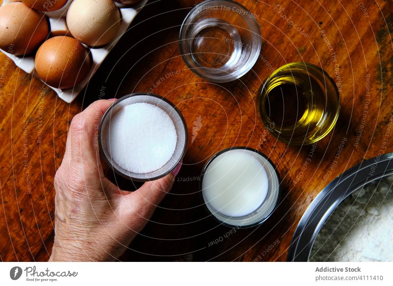 Crop woman taking pastry ingredient from table cook kitchen home cup donut food female sugar oil water milk dairy egg flour prepare raw gourmet cuisine fresh