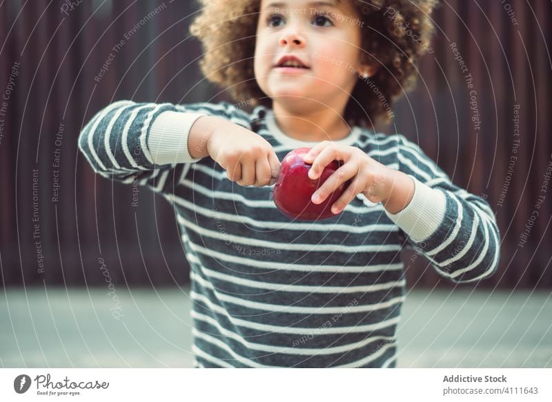 Funny little girl eating apple on street kid funny style snack happy grimace fruit trendy cute curly hair child adorable smile casual cheerful fashion outfit