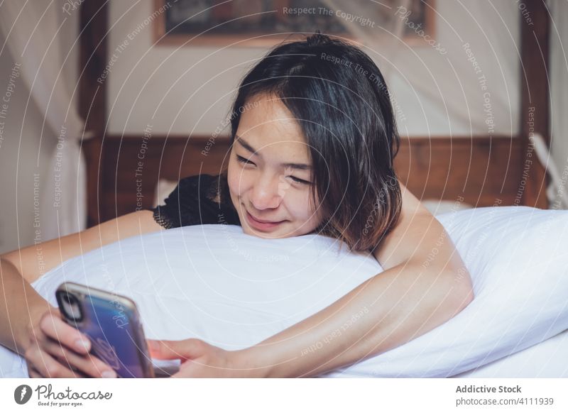 Happy Asian woman using smartphone in bed happy smile ethnic pillow home cozy female relax rest bedroom young comfort device gadget browsing watch lifestyle