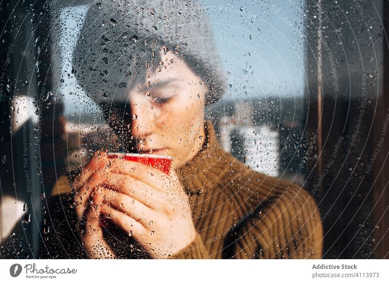 Thoughtful woman drinking coffee near window sad depression self isolation home unhappy lonely covid19 melancholy young female wet rain solitude stress cup