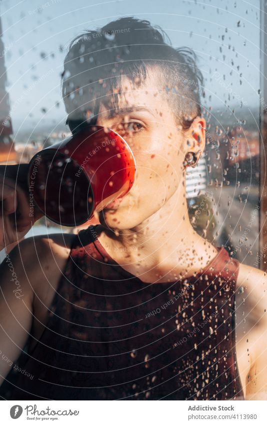 Thoughtful woman drinking coffee near window sad depression self isolation home unhappy lonely covid19 melancholy young female wet rain solitude stress cup