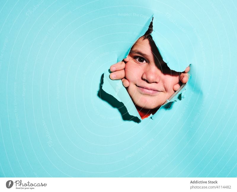 Happy boy looking through paper hole kid grimace ripped expressive concept child male copy space looking at camera blue background personality plus size