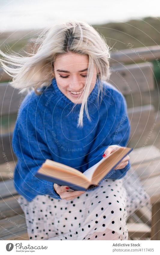 Young woman reading book on seashore terrace bench beach rest young blue sweater nature relax female wooden style vacation enjoy holiday modern trendy blond