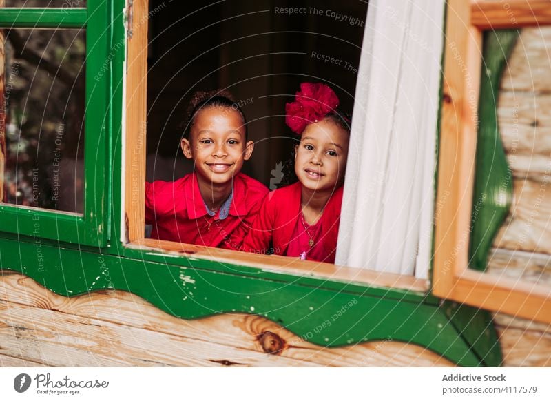 Cheerful ethnic children looking out window kid house happy sibling together cheerful wooden rural smile home lifestyle countryside joy little casual sister