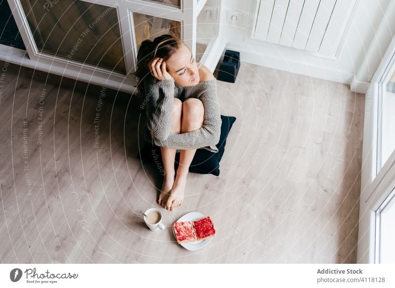 Dreamy woman having breakfast near window at home dream pensive floor toast coffee casual relax morning alone sit young female lifestyle rest serene tranquil