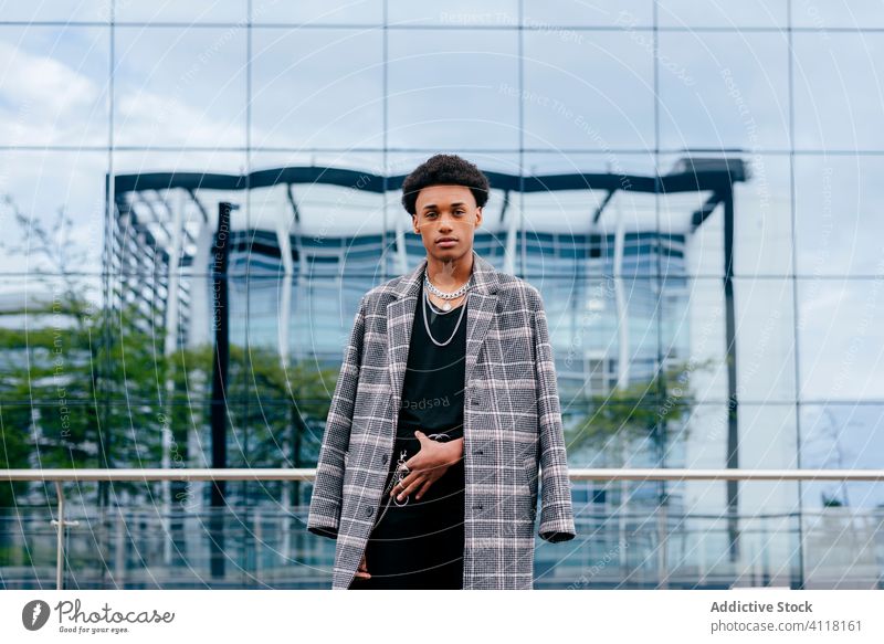 Stylish ethnic man in coat against glass wall trendy fashion urban street style chain accessory model city building serious modern african american black