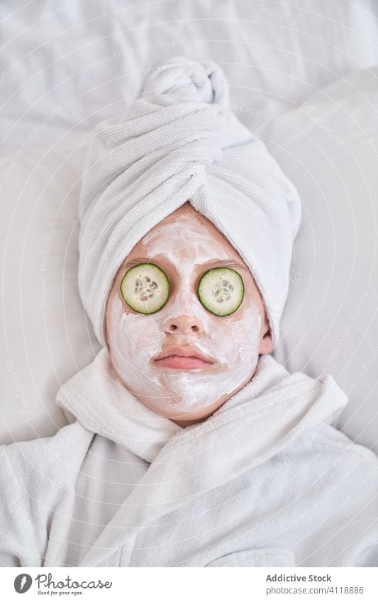 Playful kid with facial mask lying on bed having fun cucumber beauty skin care grimace playful show tongue enjoy spa joke humor natural child happy make face