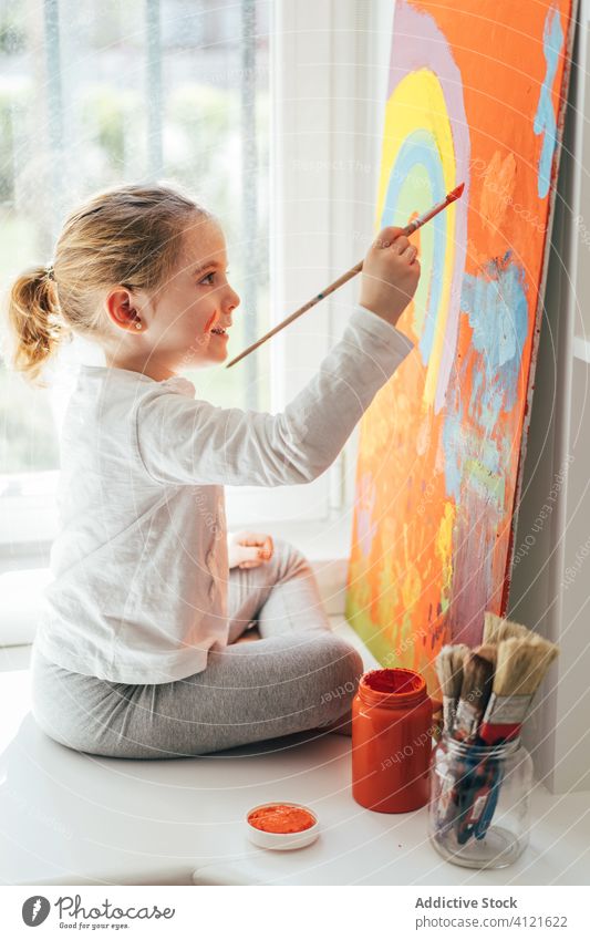 Little girl painting picture with paintbrush canvas draw rainbow creative colorful orange sill casual window art hobby imagination bright sit indoors