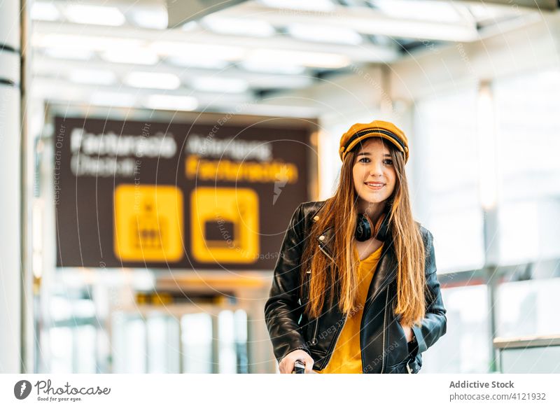 Trendy young woman in modern airport terminal tourist trendy passenger cheerful headphones style fashion departure smile trip handle wait black leather jacket