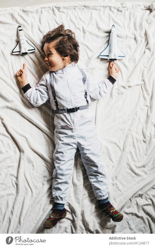 Boy in astronaut costume lying on bed boy spacesuit toy spaceship having fun smile kid cosmonaut bedroom child cheerful weekend rocket playful uniform pretend