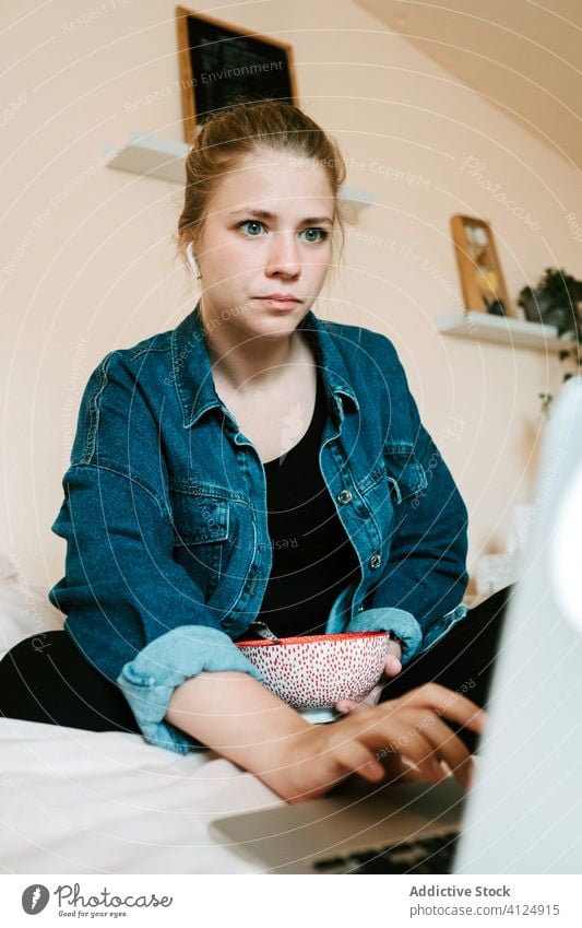 Shocked young woman in earbuds using laptop while having lunch in light modern bedroom at home shock eat earphones read watch stun social media film movie