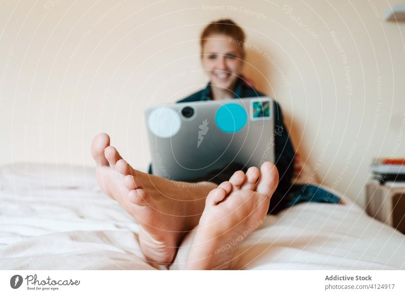 Happy young woman using laptop with pleasure while chilling in light bedroom staying at home use relax foot happy barefoot social media watch browsing surfing