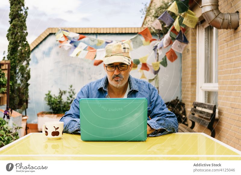 Pensive senior man in glasses having hot drink and using laptop on terrace of country house pensive read watch mug thoughtful device coffee internet male