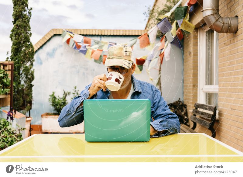 Pensive senior man in glasses having hot drink and using laptop on terrace of country house pensive read watch mug thoughtful device coffee internet male