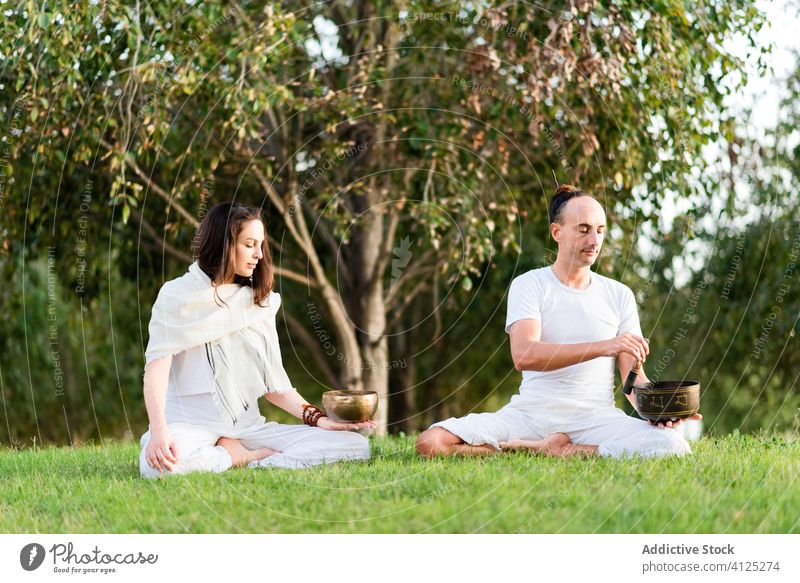 Concentrated couple meditating while sitting in lotus position yoga singing bowl tibetan bowl flexible lotus pose meditate calm eyes closed zen lawn enjoy green