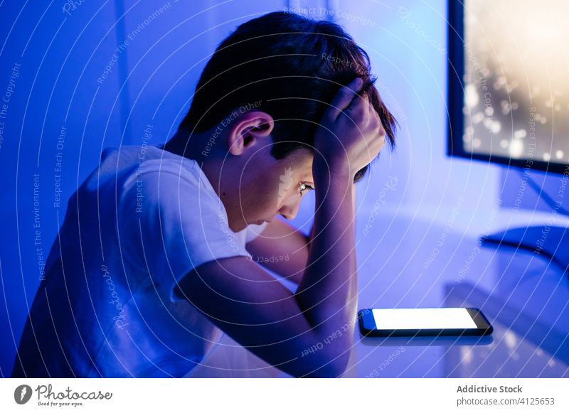 Concerned male child watching smartphone screen while sitting at table boy touch head confused empty screen bewildered surfing internet using gadget device