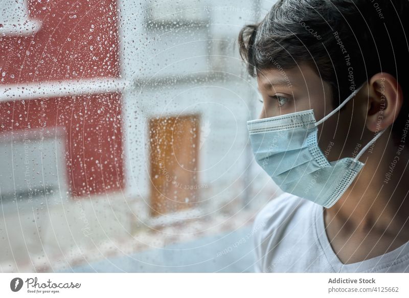 Sad boy in medical mask near window coronavirus epidemic home sad unhappy self isolation social distancing child kid face mask stand lean observe rain street