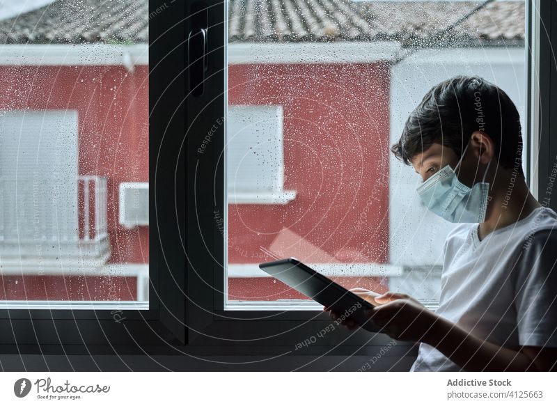 Boy in medical mask using tablet during self isolation boy quarantine kid coronavirus browsing child home epidemic social distancing stand window face mask rain