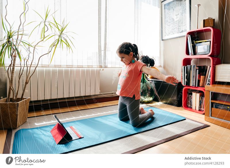 Joyful little girl doing yoga in light room kid pose mat video tablet tutorial balance practice at home stress relief watch harmony stretch flexible spirit