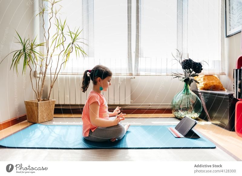 Barefoot girl doing meditation in cozy room at home kid yoga lotus pose concentrate zen spirit meditate tablet tutorial video mat peace balance practice